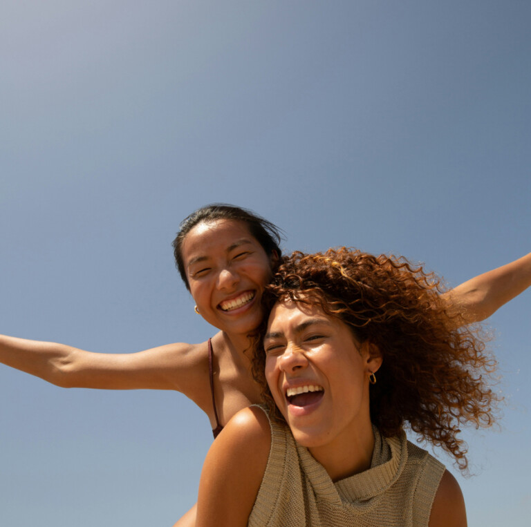 Two smiling women laughing with their arms outstretched against a clear blue sky, capturing the joy of a personalised property marketing campaign adapting in real-time.