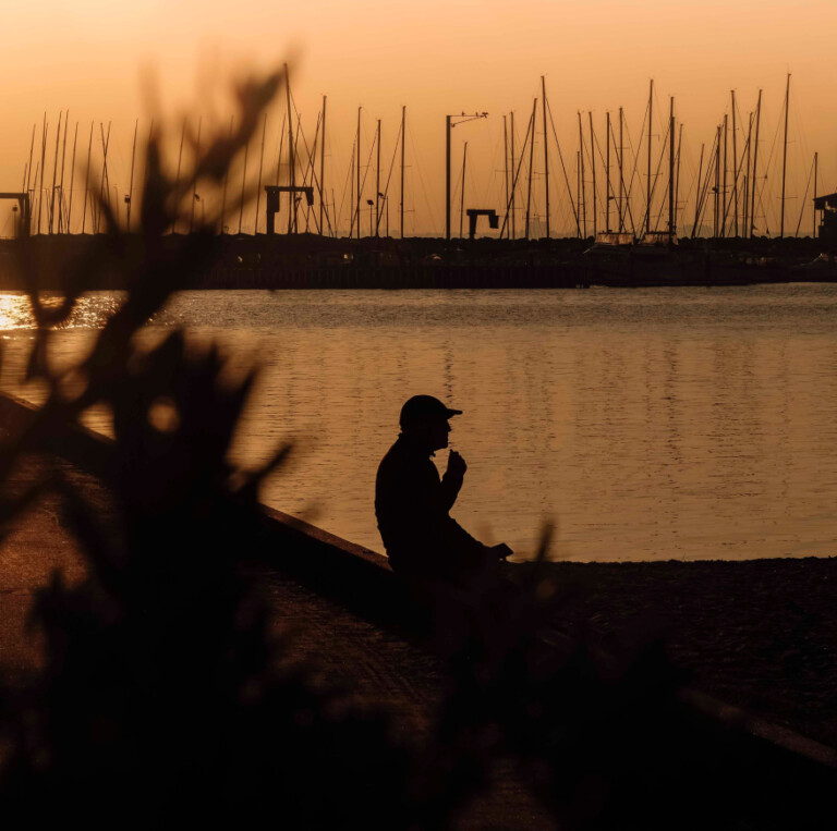 A silhouette of a person sitting by a harbour at sunset, with numerous sailboat masts visible in the distance, representing a personalised property marketing campaign adapting in real time.