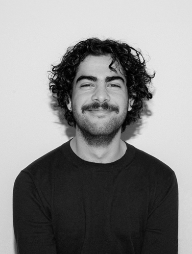 A portrait of a smiling person with curly hair and a moustache wearing a dark crew-neck shirt.
