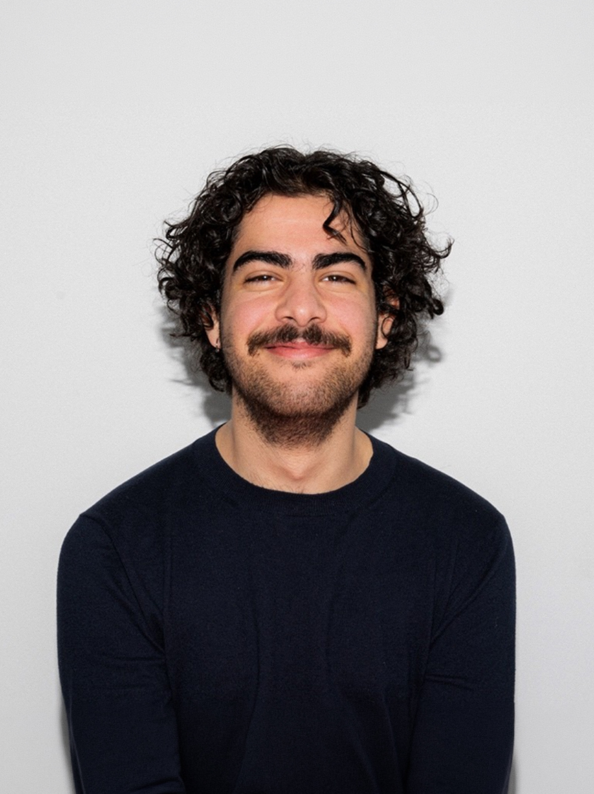 A young man with curly dark hair and a moustache smiling against a white background.