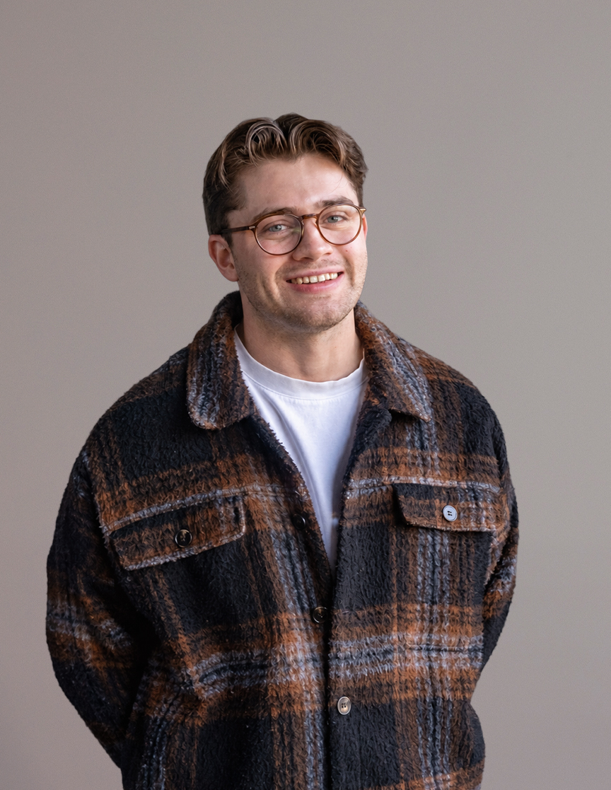 A man wearing glasses and a checked button-up jacket over a white t-shirt, smiling at the camera.